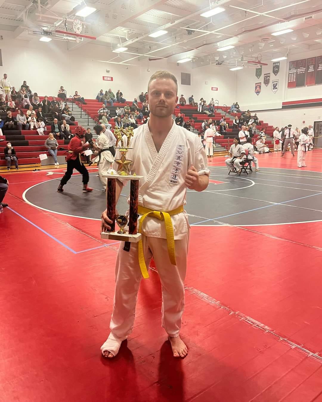 Karate competitor posing with a trophy beside tournament mats